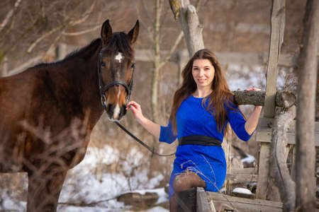 A beautiful girl is sitting on a log against the background of wooden ruins, a horse is standing nearbyの写真素材