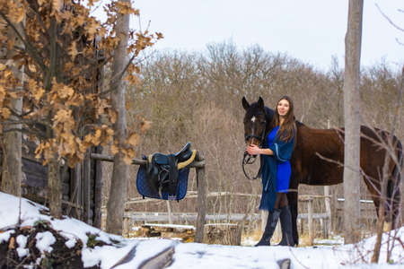 A girl in a blue dress walks with a horse through a farm in winterの写真素材
