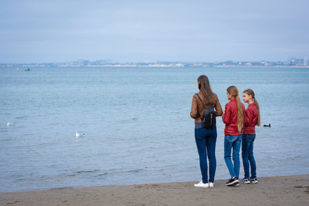 Mom and two daughters stand on the seashore on an autumn day, rear view