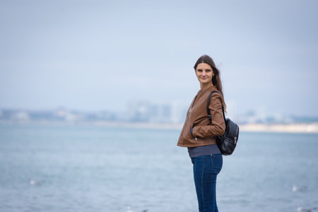 A girl in autumn clothes with a backpack stands on the seashoreの写真素材