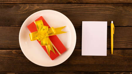 gift in a red box with a yellow bow and a white postcard note on a wooden background. gift in a white plateの写真素材