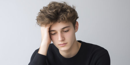 Young man with curly hair, wearing a black sweater, is thoughtfully resting his head on his hand, conveying a sense of contemplation and introspection in a minimalistic settingの素材