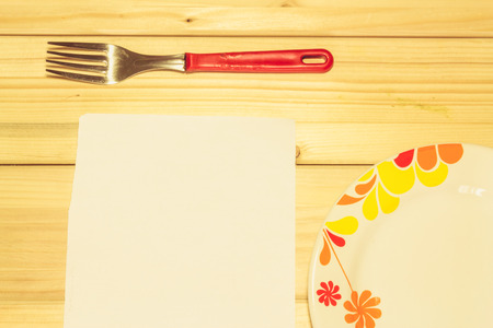 Close up shot to an empty white plate with floral design next to a fork and a piece of a white plain paper on a wooden backgroundの写真素材