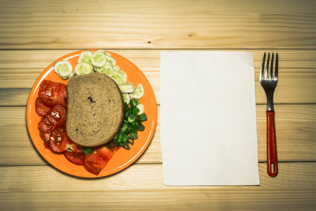 A few sliced vegetables on a plate with aslice of bread on top of them next to a white plain piece of paper and a fork on a wooden backgroundの写真素材
