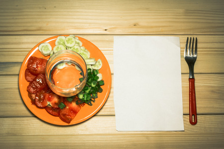 A few sliced vegetables on a plate with an empty glass on top of them next to a white plain piece of paper and a fork on a wooden backgroundの写真素材