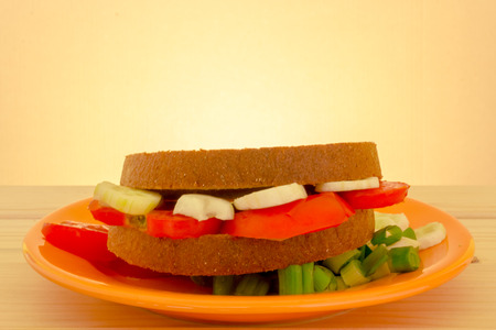 Shot to a vegetarian sandwich on a plate full of tomato slices and a few green onion pieces on a wooden table with a light colored backgroundの写真素材
