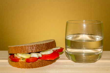 Shot to a vegetarian sandwich made from a few slices of tomato and a few slices of cucumber next to a glass of water on a wooden tableの写真素材