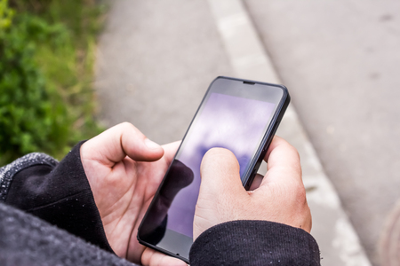 Close up shot to a guy's hands holding and taping a mobile phone while standing on a sidewalkの写真素材