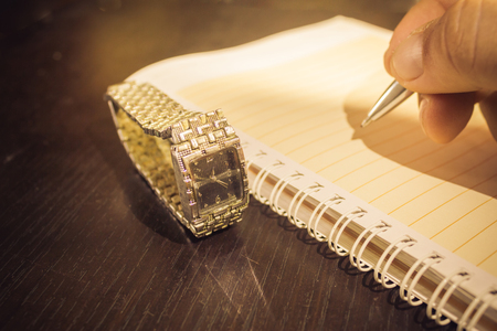 Close up shot to an agenda, a hand holding a pen and an vintage watch on a dark wooden tableの写真素材