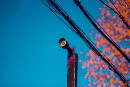 Low angle closeup of a single black surveillance camera with a blue sky in the background and a line of black wiresの写真素材