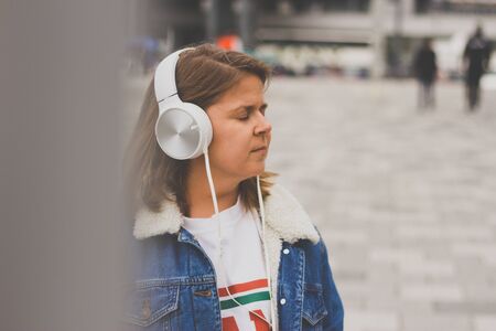 Young woman holding her eyes closed and enjoying music to a big white pair of headphones â Cute lady wearing a denim jacket and bright headsets in an urban scapeの写真素材
