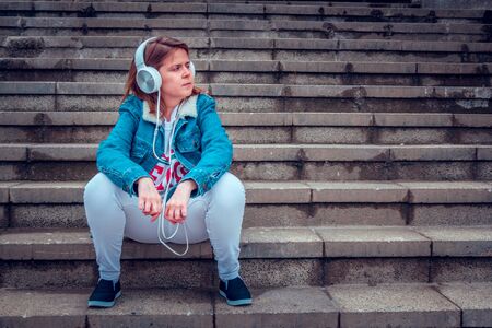 Cute lady wearing a denim jacket and a pair of big white headphones enjoying music in the city â Young woman sitting on concrete stairs with hands leaning on her legs and looking away in the cityの写真素材