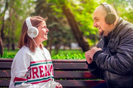 Boy wearing a black leather jacket and girl with a white blouse enjoying music at their big headphones â Young man and woman sitting on a bench outdoor in nature looking at each other one listening energic tune and the other a soft oneの写真素材