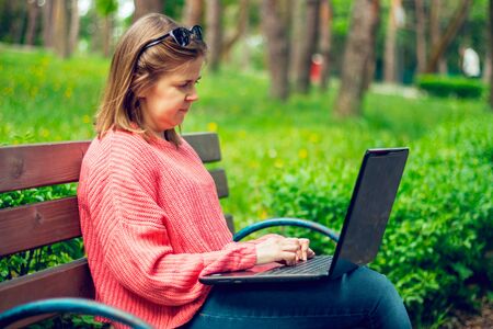 Young woman wearing a pink blouse and jeans with laptop in the park â Girl sitting on a bench outdoors holding and using her computer â Female student working on a notebook in natureの写真素材