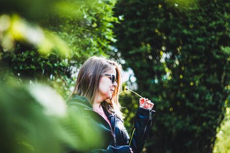 Young woman with sunglasses and a leather jacket smelling a small dandelion â Girl holding a little yellow flower on a bright spring day with trees in the backgroundの写真素材