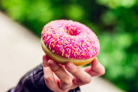 Closeup of a pink glazed doughnut with colored sprinkles on top hold by a personâs hand in the park - Tasty and delicious fresh baked treat for desert â Concept image for street foodの写真素材