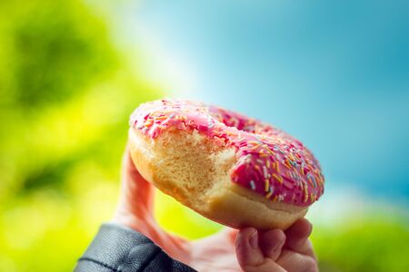 Hand holding a pink glazed donut with colorful sprinkles on top in the park â Tasty baked desert for lunch break â Concept image for street food and junk foodの写真素材
