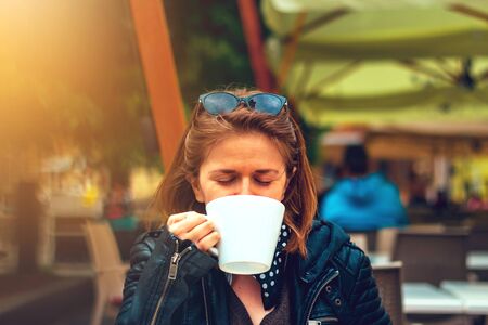 Young woman with brown hair wearing sunglasses a leather jacket and a scarf with dots â Casually dressed millennial holding and drinking a hot beverage from a big white mugの写真素材