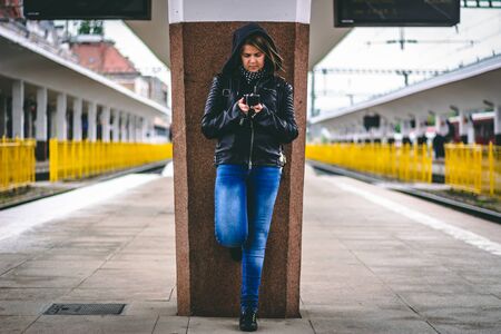 Woman wearing jeans and a black leather jacket using the phone in a station â Casually dressed millennial texting on a mobile phone while leaning on a pillar with one leg upの写真素材