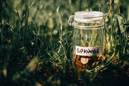 Small glass jar with golden coins with the label savings sitting in the grass on a bright spring day  Concept image for planning money, saving, achieving goals and lifestyleの写真素材