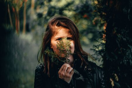 Young woman with brown hair holding a small green thuja branch in front of her face on a bright...