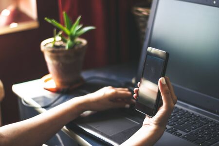 Woman hand holding a big black smartphone while working on a laptop keyboard with a green plant in background indoor during day time â Concept image for multitasking and online shopping, perfume,の写真素材