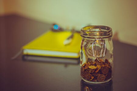 Small closed jar half full of golden coins with a green notebook and a pair of glasses in background sitting on a desk indoor â Concept image for saving and planning moneyの写真素材