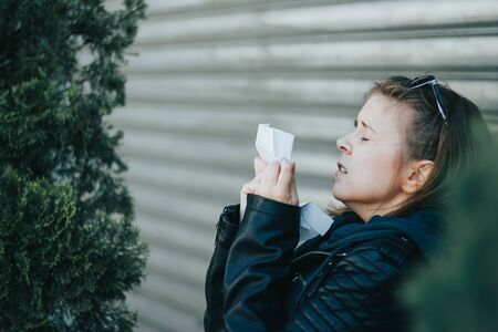 Young woman with brown hair wearing leather jacket and sunglasses sneezing in a white napkin with her eyes closed outdoor â Girl having an allergic reaction or a coldの写真素材