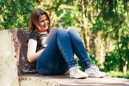 Happy young woman with short brown hair wearing a black t-shirt and jeans sitting down in a park and reading a book on a sunny spring day â Concept image for enjoying literature anywhereの写真素材