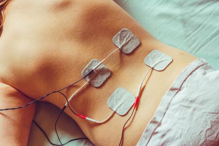 Small white electrodes placed on a womanâs back laying down on a white sheet â Electric device with tens and ems alternative current used for pain relief and massageの写真素材
