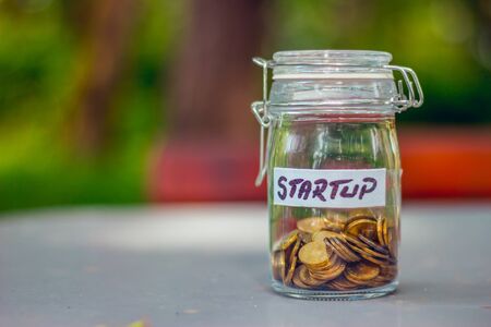 Closed small glass jar with coins in it and a label with the word startup written on it on a table with a green blurry background â Concept image for investment, a new start or raising foundsの写真素材