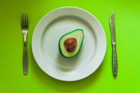 Half of green and ripe avocado with seed sitting on a white large plate with knife and fork on aside â Minimalistic dish from a vegan diet full of oils and nutrientsの写真素材