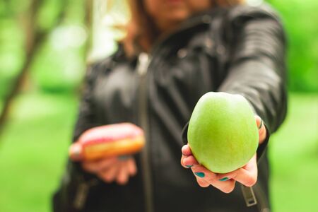 Hand holding a green apple in one hand and pink glazed doughnut in the other in nature â Girl giving healthy and delicious alternative for desert â Woman choosing fruit instead of caloric sweetの写真素材