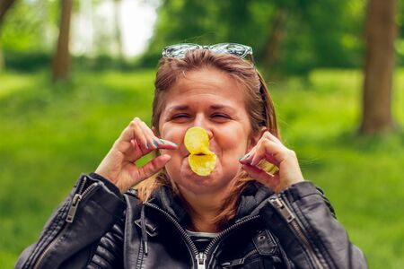 Playful young woman wearing leather jacket, making a duck beak from potato chips in the park â Pretty grill feeling god and joyful playing with snacksの写真素材