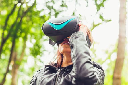 Young woman wearing a leather jacket using a pair of virtual reality glasses in nature â€“ Girl...