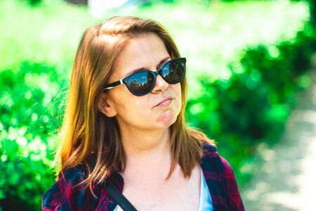 Smiling young woman with short brown hair wearing black sunglasses standing in the bright sunlight in nature â Casually dressed girl enjoying the summer heat outdoorの写真素材