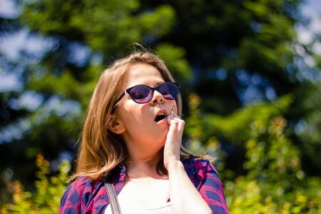 Young woman with short brown hair wearing sunglasses having a toothache outdoor â Girl sitting in the bright sun while experiencing a dental pain and holding her hand on her jawの写真素材