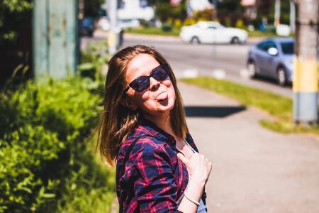 Young woman with short brown hair wearing a plaid shirt and sunglasses having fun in the city â Casually dressed millennial anting silly while sticking out her tongueの写真素材