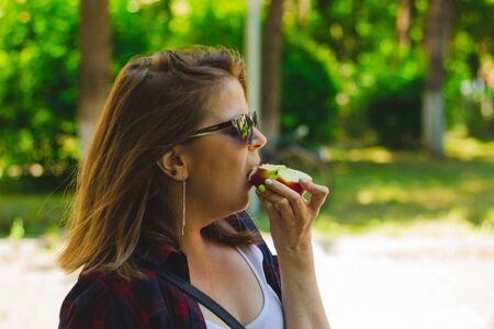 Young woman with short brown hair wearing sunglasses biting from a red apple while walking in nature...