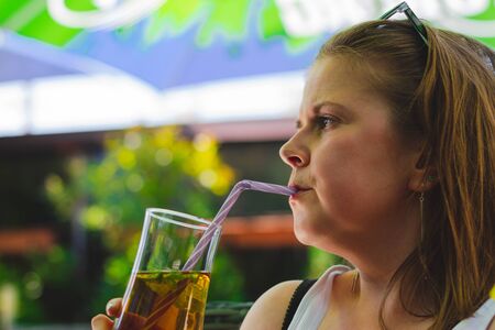 Young woman with short brown hair drinking a cold ice tea from a glass with a straw on a hot day â Girl sitting on a terrace and sipping a iced fruity refermentの写真素材