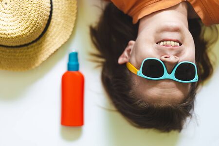 Happy young woman with colorful sunglasses lying next to yellow straw hat and sun lotion on a white background â Smiling cute girl with basic summer accessoriesの写真素材
