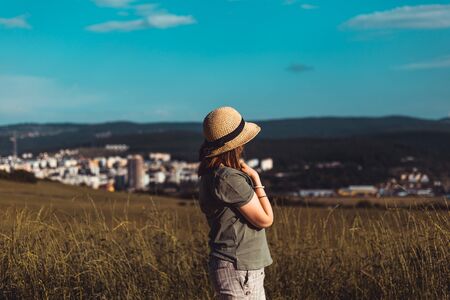 Young woman wearing summer outfit relaxing while looking at cityscape from a high green hill -...