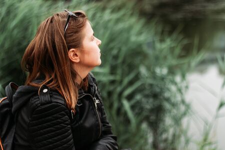 Young woman with short brown hair daydreaming while sitting near a lake in a park â Pretty girl wearing a leather jacket enjoying peaceful moments in a natural environmentの写真素材
