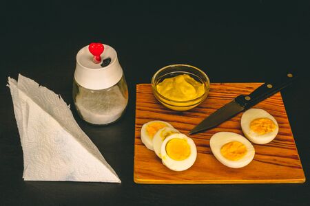 Eggs served as breakfast on dark background â Organic lunch of yolks and a bowl of mustard next to a kitchen knife, a salt shaker and a napkinの写真素材