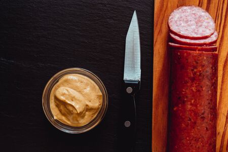 Salami with slices next to a kitchen knife and a bowl of mustard â Traditional chopped pork meat recipe with seasoningの写真素材