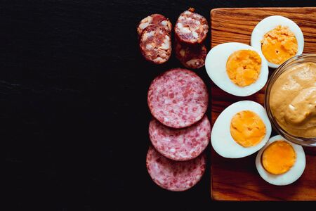Healthy and fresh organic food served as appetizer with copy space - Hardboiled egg halves and slices of smoked salami and sausage on black stone textured backgroundの写真素材
