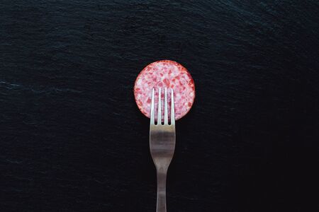Smoked salami slice with fork on top placed on black background â Spicy traditional homemade meat appetizer on stone textured surfaceの写真素材