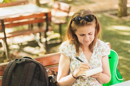Young woman taking notes while sitting on a bench in the park on a sunny day - Pretty girl with brown hair handwriting in a small notebook - Female student writing information outdoorsの写真素材