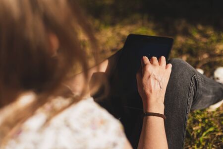 Young woman using a tablet outdoors on a sunny day - Millennial girl with brown hair holding a wireless modern gadget while sitting in the grass in a parkの写真素材