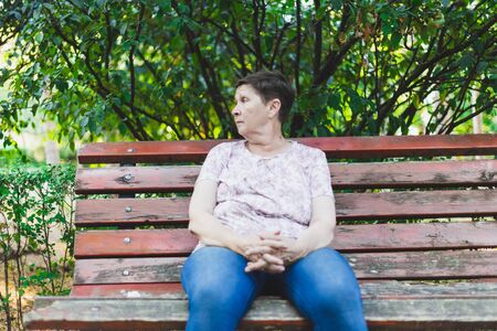 Sad senior woman sitting alone on a bench while looking in the distance - Depressed and confused cute old lady with short brown hair feeling lonely in the parkの写真素材